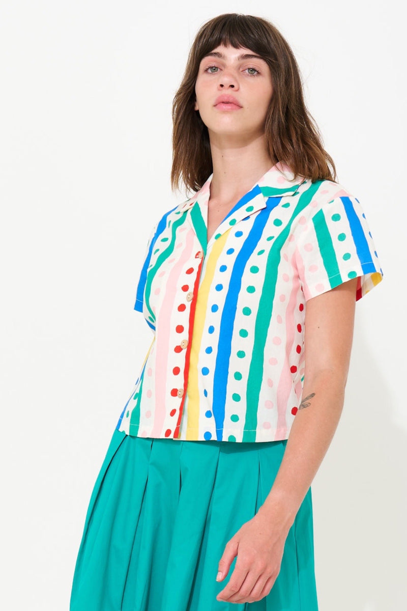 A woman wearing a short-sleeve collared button-up shirt with vertical stripes in blue, green, yellow, red, and pink on a white base, paired with a teal skirt, against a plain white background.