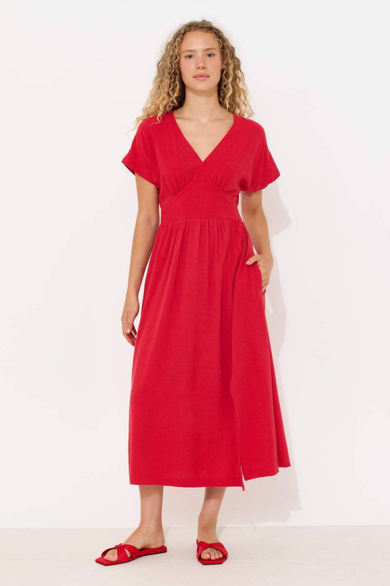 A woman modeling a bright red midi dress with short sleeves, a V-neckline, and side pockets, paired with matching red slide sandals against a plain white studio background.