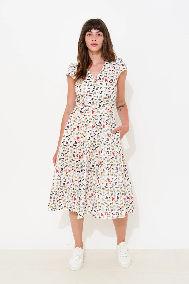 A woman modeling a white midi dress with a colorful, dense all-over print of small figures, set against a plain white studio background.