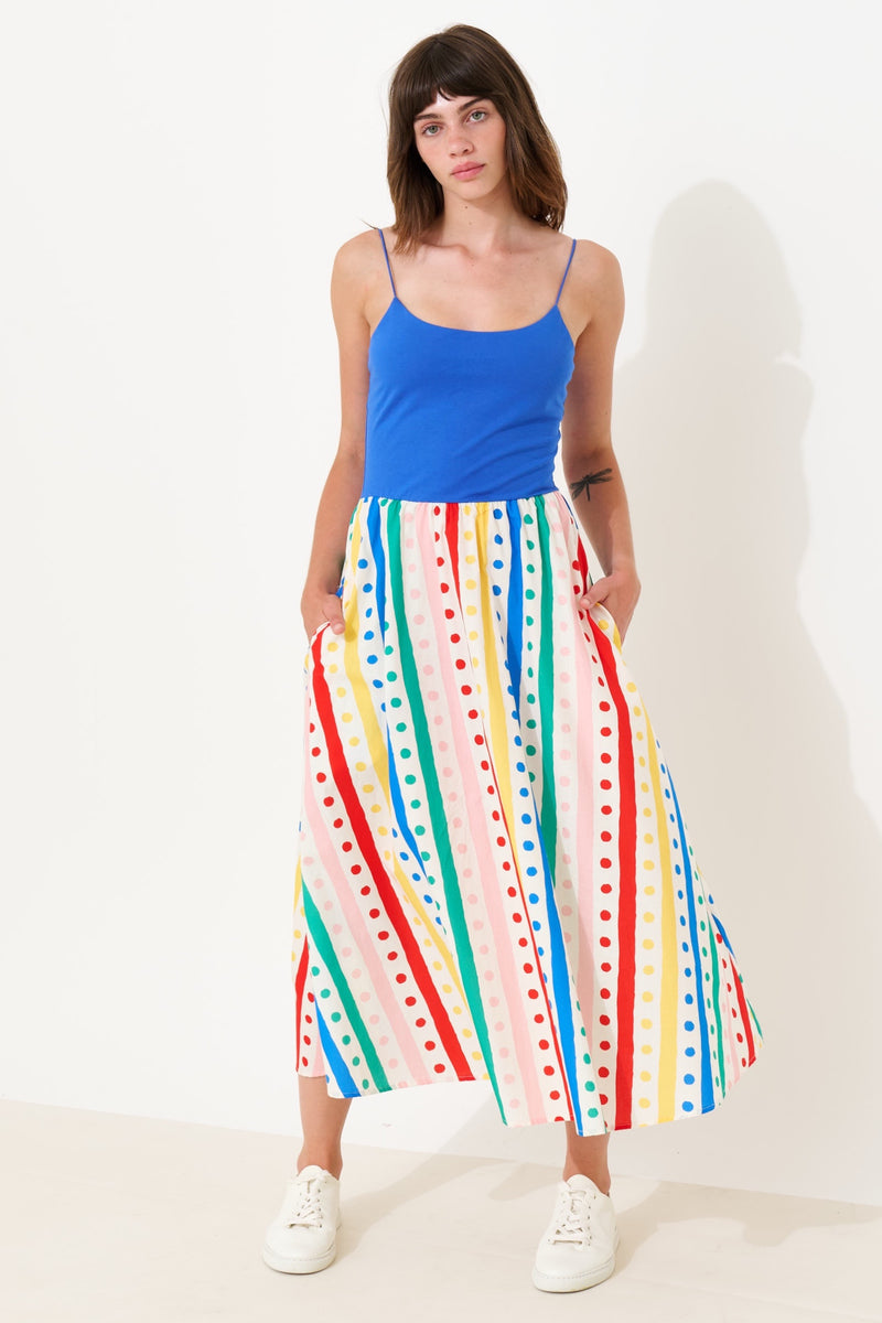 A woman modeling a bright blue camisole top and a long midi skirt with vertical multicolor stripes and polka dots in pink, blue, green, yellow, and red, paired with white sneakers against a plain white studio background.