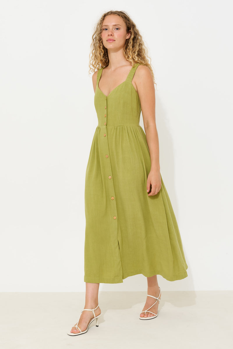 A woman modeling a sleeveless olive green midi dress with a button-down front and thin straps, paired with white strappy high-heeled sandals against a plain white studio background.